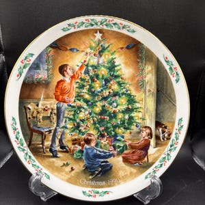 May include: A decorative plate featuring a Christmas scene with a decorated tree, children, and a dog. The plate has a white border with holly and berry accents. The text "Christmas 1990" is at the bottom.