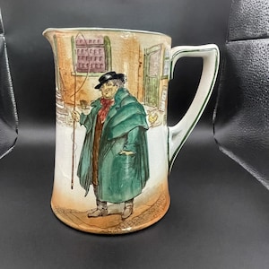 Puede incluir: Un jarro de cerámica vintage con asa, con una ilustración pintada a mano de un hombre con abrigo y sombrero verdes. La obra de arte representa una escena callejera con edificios y una figura que sostiene un bastón. El jarro tiene una base blanca con un diseño detallado y colorido.