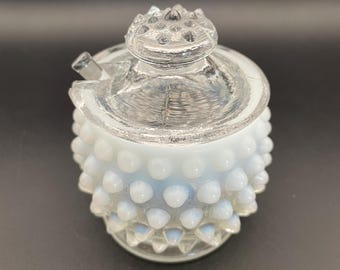 May include: A clear glass sugar bowl with a lid and a small spout. The bowl has a white, hobnail-like texture on the lower portion. The lid is clear with a textured top. The sugar bowl is on a black background.