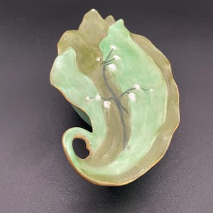 May include: A leaf-shaped ceramic dish in light green and olive green. It has a gold-coloured rim and a painted floral design with white accents. The dish is approximately 15 cm long.