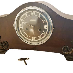 May include: A dark brown wooden mantel clock with a rounded top, a circular face featuring white Arabic numerals, and a silver-toned bezel. The clock is decorated with gold-colored ornate scrollwork on the lower corners and includes a metal winding key.