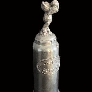 Peut inclure: Un récipient en métal argenté avec un couvercle, orné d'une figurine d'ours décorative tenant une boule. Le récipient porte une étiquette ovale avec l'inscription "PICNIC BUBBLES". L'objet est posé sur un fond noir.