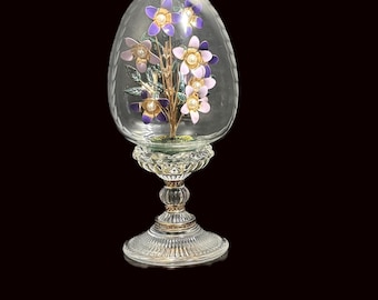 May include: A clear glass egg-shaped decorative piece with a floral arrangement inside. The flowers are purple and pink with gold accents and pearl centers. The egg sits on a clear, ornate glass pedestal.