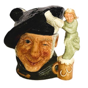 May include: A ceramic character jug featuring a winking person wearing a large black hat and dark clothing. The handle is shaped like a smaller figure in a green outfit standing atop a brown mug decorated with two white horseshoe symbols.
