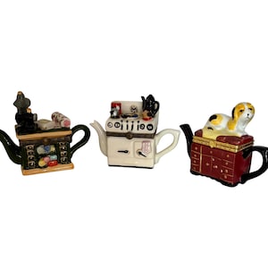 May include: Three decorative ceramic teapots shaped like miniature furniture pieces. The left is a green cabinet with a sewing machine, the centre is a white kitchen stove, and the right is a red chest of drawers topped with a small dog figurine.