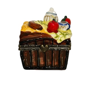 May include: A brown ceramic trinket box shaped like a picnic basket with a hinged lid. The lid features a painted yellow and white checkered cloth, a white teapot, a red apple, and a small blue and white cup. A metal clasp with a heart shape.