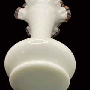 May include: A white glass vase with a round body and a ruffled rim. The rim has a delicate pinkish-orange trim. The vase sits on a circular base, and the background is black.