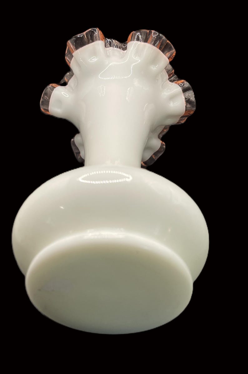 May include: A white glass vase with a round body and a ruffled rim. The rim has a delicate pinkish-orange trim. The vase sits on a circular base, and the background is black.