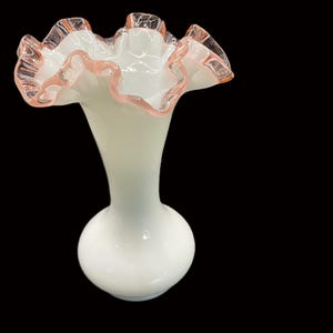 May include: A white glass vase with a ruffled rim, accented with a delicate pink trim. The vase has a bulbous base and a slender neck, creating an elegant silhouette. The ruffled edge adds a touch of vintage charm.