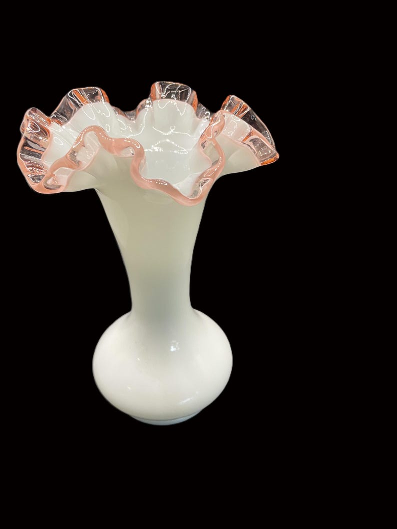 May include: A white glass vase with a ruffled rim, accented with a delicate pink trim. The vase has a bulbous base and a slender neck, creating an elegant silhouette. The ruffled edge adds a touch of vintage charm.