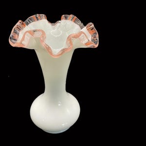 May include: A white glass vase with a ruffled rim. The rim is trimmed with a delicate pink color. The vase has a bulbous base and a slender neck, creating an elegant silhouette. The vase is likely used for displaying flowers.