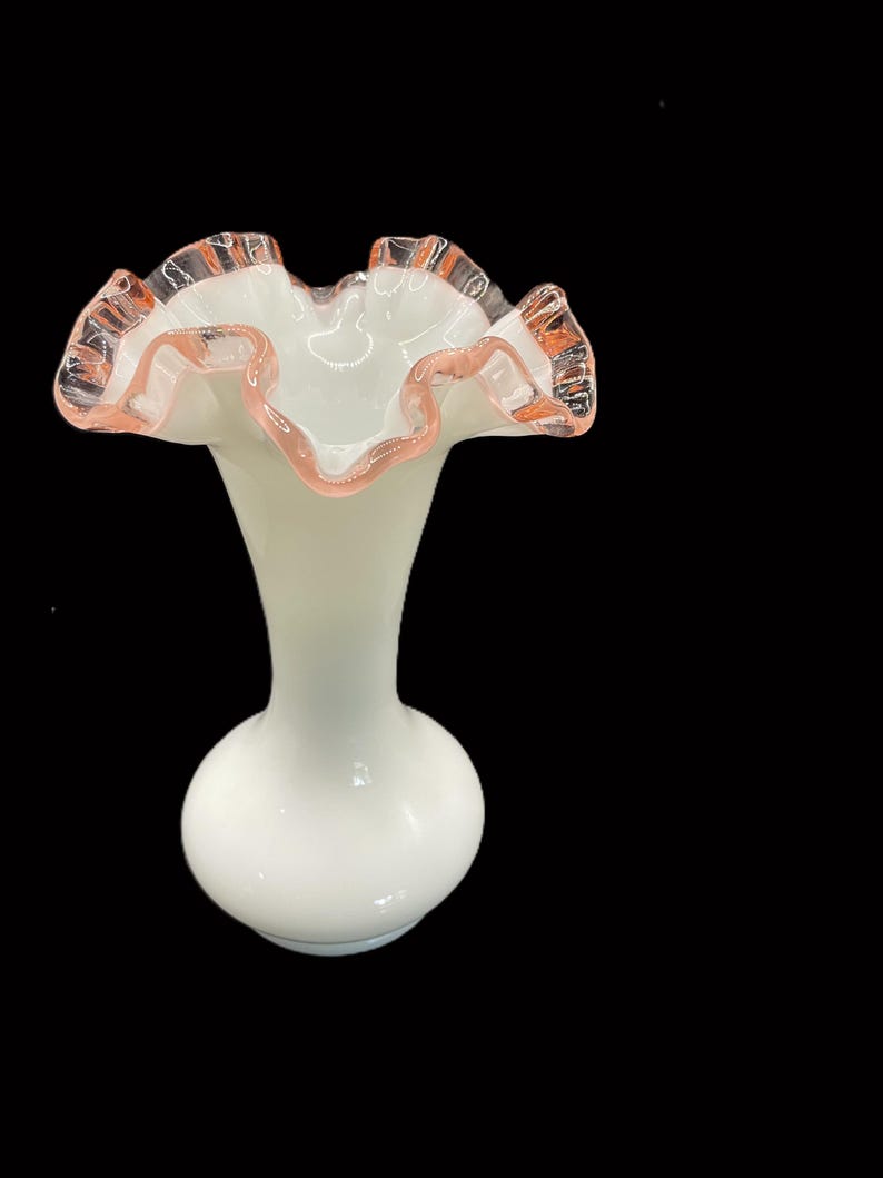 May include: A white glass vase with a ruffled rim. The rim is trimmed with a delicate pink color. The vase has a bulbous base and a slender neck, creating an elegant silhouette. The vase is likely used for displaying flowers.