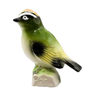 May include: A ceramic bird figurine featuring a green back, yellow wings with black tips, and a white chest. The head has a black and white striped pattern with an orange crown. The bird stands on a light base with the number 26 embossed on it.