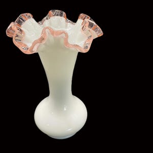 May include: A white glass vase with a ruffled top edged in pink. The vase has a bulbous base and a slender neck, creating an elegant silhouette. The ruffled edge adds a delicate, feminine touch to the design.