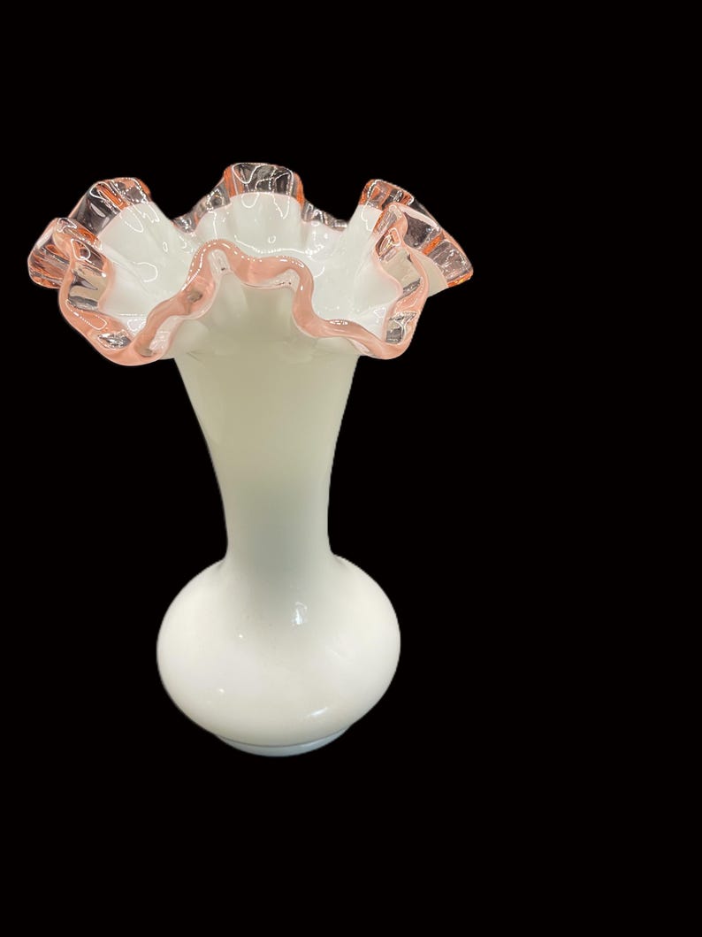 May include: A white glass vase with a ruffled top edged in pink. The vase has a bulbous base and a slender neck, creating an elegant silhouette. The ruffled edge adds a delicate, feminine touch to the design.