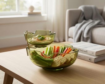 May include: A two-tiered olive green glass serving dish with a wavy rim. The top tier holds a small portion of hummus garnished with a green herb, while the larger bottom tier contains an assortment of sliced raw vegetables and round crackers.