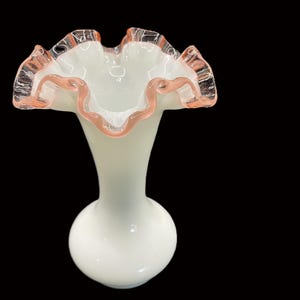 May include: A white glass vase with a ruffled rim, accented with a delicate pink trim. The vase has a bulbous base and a slender neck, creating an elegant silhouette. The ruffled edge adds a touch of vintage charm.
