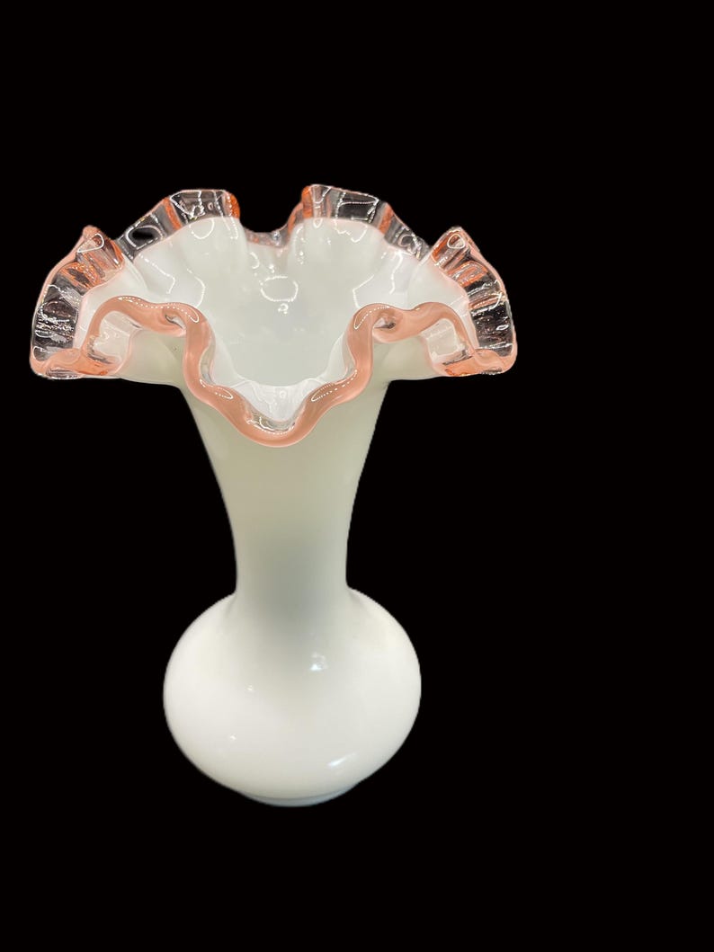 May include: A white glass vase with a ruffled rim, accented with a delicate pink trim. The vase has a bulbous base and a slender neck, creating an elegant silhouette. The ruffled edge adds a touch of vintage charm.