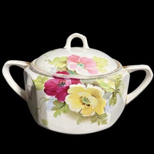 May include: A white ceramic sugar bowl with a lid, featuring hand-painted floral designs in shades of pink, yellow, and red. The bowl has two handles and a small loop handle on the lid. Gold trim accents the edges.