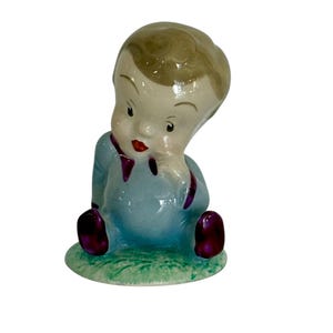 May include: A vintage ceramic figurine of a seated baby. The baby wears a light blue outfit with purple accents and shoes. The figurine has a light skin tone, blonde hair, and red lips. It sits on a green base.