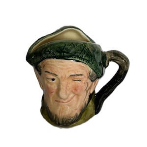 以下が含まれることがあります： ウィンクした表情の人物の頭部を模した陶器製のキャラクタージャグ。フィギュアは質感のあるダークグリーンの帽子をかぶり、明るい肌の色にバラ色の頬と濃い髭が特徴です。右側には曲線的なハンドルが付いています。