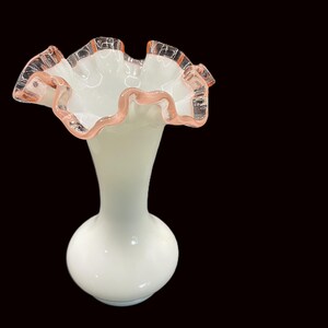 May include: A white glass vase with a ruffled rim. The rim is edged with a delicate pink trim. The vase has a bulbous base and a slender neck, creating an elegant silhouette. The background is black.