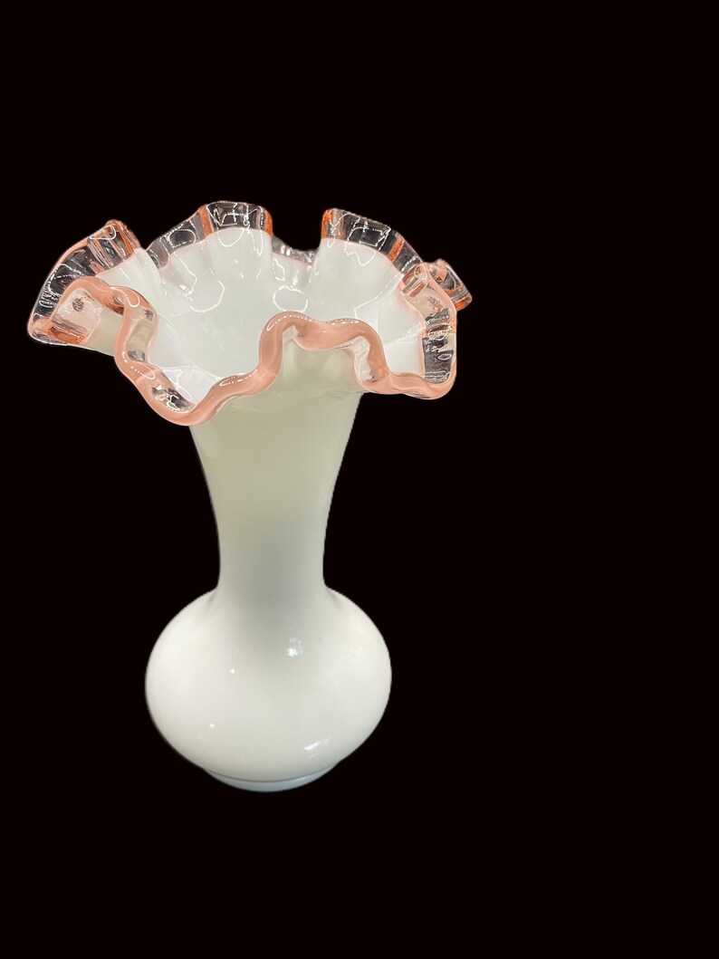 May include: A white glass vase with a ruffled rim. The rim is edged with a delicate pink trim. The vase has a bulbous base and a slender neck, creating an elegant silhouette. The background is black.