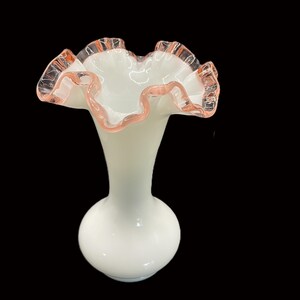 May include: A white glass vase with a ruffled rim. The rim is edged with a pink hue. The vase has a bulbous base and a slender neck, creating an elegant silhouette. The background is black.