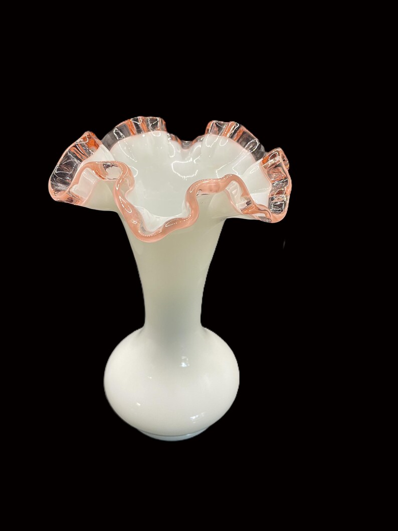 May include: A white glass vase with a ruffled rim. The rim is edged with a pink hue. The vase has a bulbous base and a slender neck, creating an elegant silhouette. The background is black.