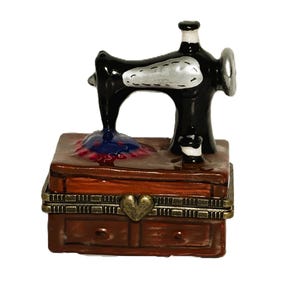 Puede incluir: Un joyero de cerámica con forma de máquina de coser antigua sobre una base marrón que imita la madera. La máquina es negra con detalles plateados y una pieza de tela azul. La base tiene una bisagra de metal cobrizo con un cierre en forma de corazón.