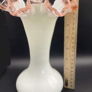 May include: A white glass vase with a ruffled rim, accented with a pink trim. The vase has a bulbous base and a slender neck. The height of the vase is approximately 10 inches.
