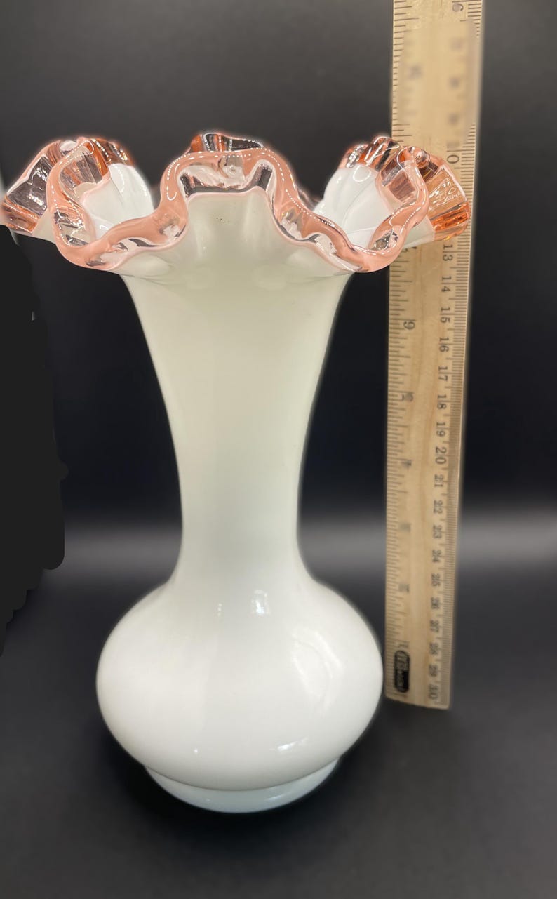 May include: A white glass vase with a ruffled rim, accented with a pink trim. The vase has a bulbous base and a slender neck. The height of the vase is approximately 10 inches.