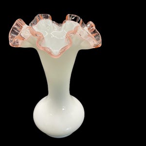May include: A white glass vase with a ruffled rim, accented with a delicate pink trim. The vase has a bulbous base and a slender neck, creating an elegant silhouette. The overall design is vintage and decorative.