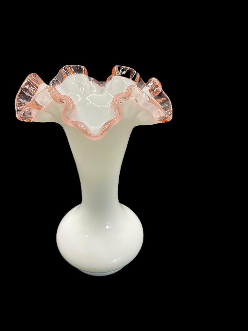 May include: A white glass vase with a ruffled rim, accented with a delicate pink trim. The vase has a bulbous base and a slender neck, creating an elegant silhouette. The overall design is vintage and decorative.