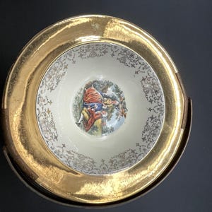 May include: A vintage porcelain bowl and plate set. The bowl features a central image of a couple in a garden scene, surrounded by a cream-coloured background with gold filigree. The plate has a wide gold rim.