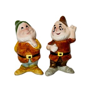 May include: Two ceramic gnome figurines standing side by side. The left gnome wears a green hat, yellow jacket, and brown belt. The right gnome wears a brown hat, brown jacket, and black belt with a yellow buckle, with one hand raised.