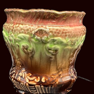 May include: A decorative ceramic planter with a textured, ruffled rim. The planter features a color palette of brown, green, and red, with embossed floral and leaf patterns. The base has a detailed, ornate design.