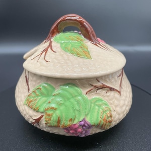 Peut inclure: Un pot en céramique beige avec un couvercle et une poignée. Le pot est décoré de feuilles vertes, de raisins violets et de vignes brunes. Le couvercle a une poignée et est également décoré de feuilles et de vignes. Le pot a une surface texturée.