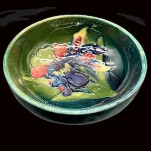 May include: A green ceramic bowl with a floral design. The interior features a raised relief of colourful flowers in shades of red, blue, and yellow against green leaves. The bowl has a glossy finish and a dark background.