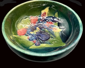 May include: A green ceramic bowl with a floral design. The interior features a raised relief of colourful flowers in shades of red, blue, and yellow against green leaves. The bowl has a glossy finish and a dark background.