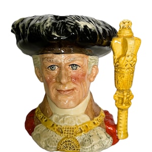 Peut inclure: Une cruche en céramique représentant une personne portant un chapeau noir à plumes et une chaîne de fonction dorée. L'anse est en forme de masse jaune. La figure a un teint hâlé, des yeux clairs et porte un vêtement rouge avec un col en dentelle blanche.