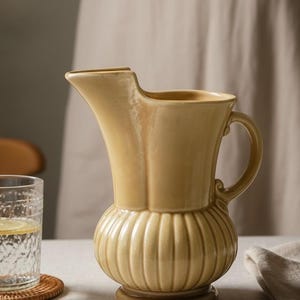 Könnte beinhalten: Ein blassgelber Keramikkrug mit geripptem, bauchigem Unterteil und glattem, ausgestelltem Oberteil, ausgestattet mit einem geschwungenen Henkel und einem ausgeprägten Ausguss, neben einem klaren Glas mit Wasser und einer Zitronenscheibe.