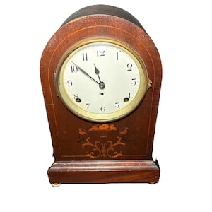 Op de afbeelding: Een vintage houten mantelklok met een ronde wijzerplaat. De klok heeft een donkerbruine afwerking met decoratieve inleg en een goudkleurige wijzerplaat. De wijzers van de klok wijzen naar ongeveer 11:58 uur.