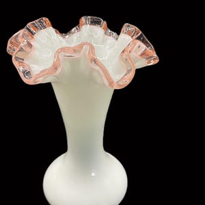 May include: A white glass vase with a ruffled rim. The rim is edged with a delicate pink color. The vase has a bulbous base and a slender neck, creating an elegant silhouette. The background is black.