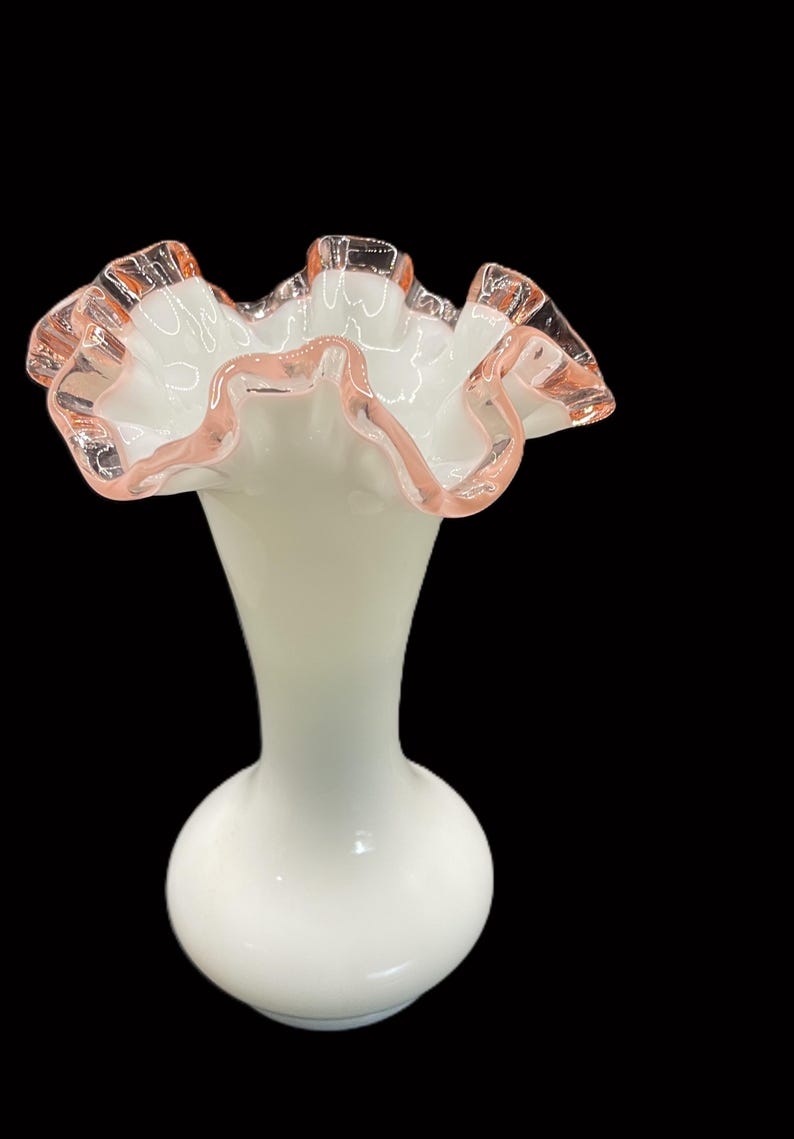 May include: A white glass vase with a ruffled rim. The rim is edged with a delicate pink color. The vase has a bulbous base and a slender neck, creating an elegant silhouette. The background is black.