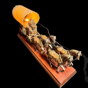 May include: A vintage lamp featuring a covered wagon pulled by four horses. The wagon has a seated figure and a lampshade. The lamp is made of bronze-colored metal and sits on a wooden base. The lamp is lit, casting a warm glow.