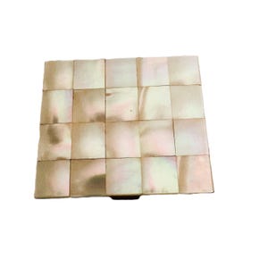 Può includere: Una scatola quadrata caratterizzata da un motivo a mosaico di tessere in madreperla iridescente. La superficie è composta da venti singoli pezzi rettangolari disposti a griglia, che mostrano tonalità bianco tenue, crema e rosa pallido con una lucentezza naturale.