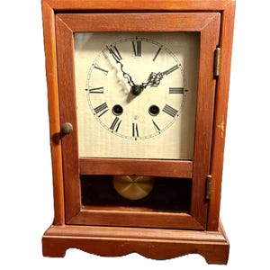 May include: A wooden antique clock with a glass front, featuring a cream-colored face with black Roman numerals and hands. The clock has a pendulum and a key is included.