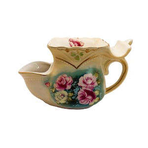 May include: A cream-colored porcelain shaving mug featuring a teal-tinted base decorated with pink and white rose floral patterns. The mug has a gold-trimmed handle, a wide opening, and a curved side rest, with a single rose painted inside the rim.