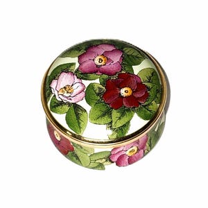 May include: A round, lidded trinket box featuring a botanical illustration of pink, dark red, and light purple flowers with green leaves on a white background. The lid and base are rimmed with a thin, polished gold-tone metal band.